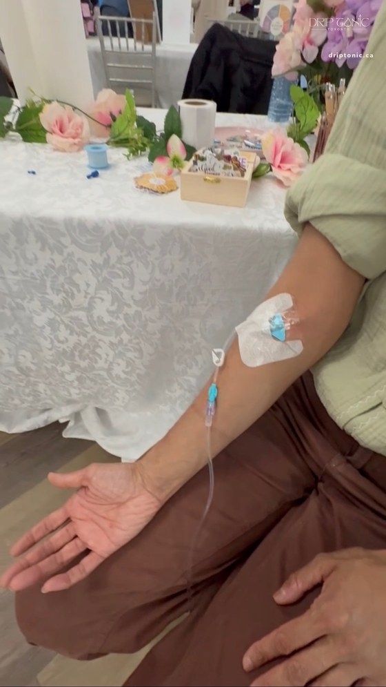 A guest at the Empower Her event receives an IV drip at the Drip Tonic Recharge Bar, her arm resting beside a floral table.