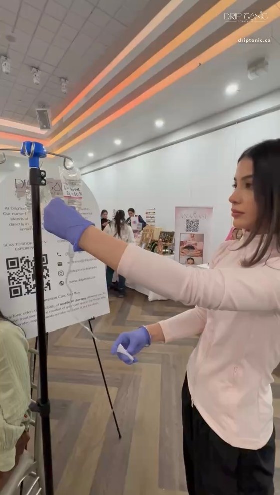 A Drip Tonic RN in gloves hangs an IV bag on the stand beside the brand's poster at the Empower Her event.