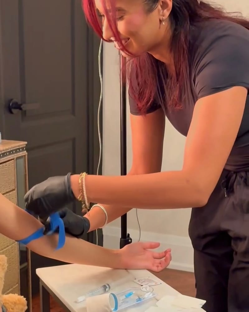 A smiling Drip Tonic RN ties a tourniquet and preps a guest's forearm for an IV cannula, the single-use setup laid out on a table at an at-home session in the GTA.