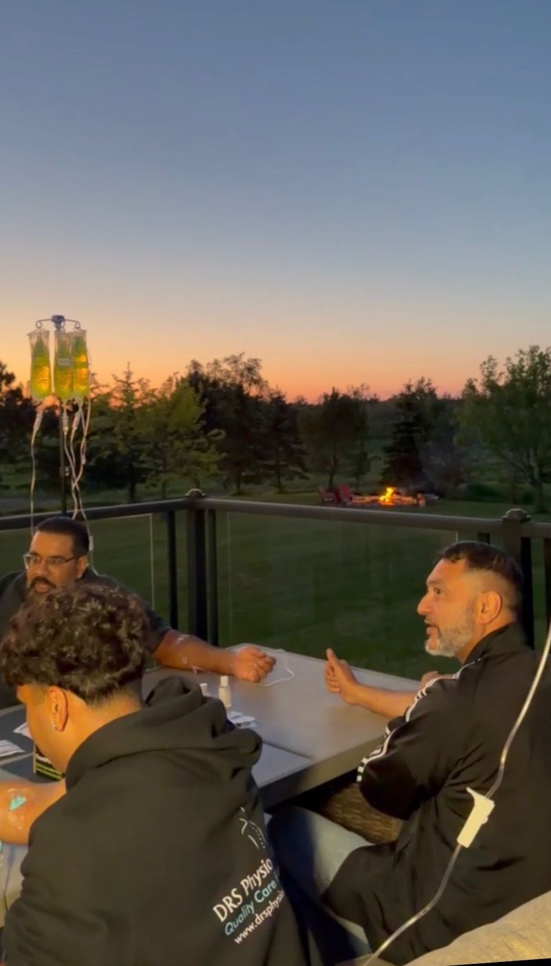Three guests receive IV drips together on a backyard deck at dusk during a Drip Tonic mobile group session near Toronto, a firepit glowing behind.