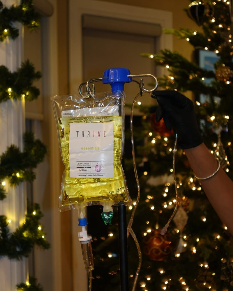 A Drip Tonic RN's gloved hand adjusts an IV drip bag on a stand beside a lit Christmas tree, an at-home holiday session in the GTA.