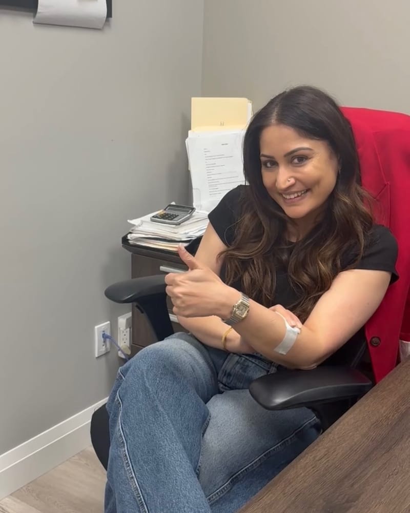 A smiling guest gives a thumbs up from a desk chair with a taped IV cannula in her forearm during a Drip Tonic office IV drip in the GTA.